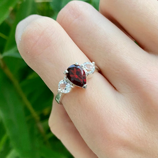 A sterling silver ring featuring a red garnet centerpiece with white topaz accents on the band, held on a hand with a blurred green background.