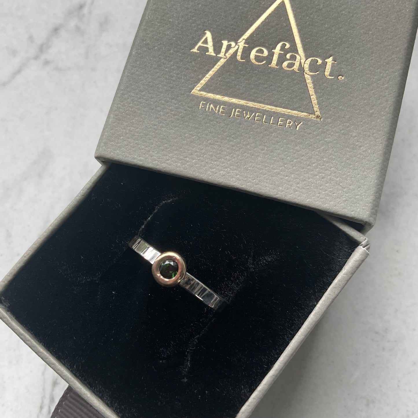 Silver ring with a gemstone in a 'Artefact' jewelry box on a light gray background