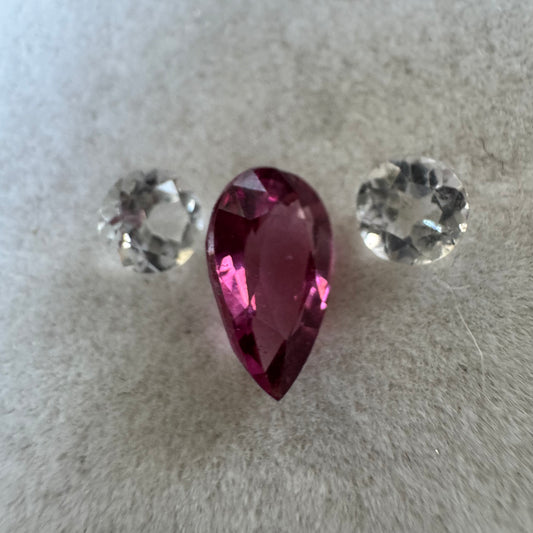 A faceted pear-shaped pink gemstone centered on a soft light gray textured surface, with two round clear gemstones placed on either side.