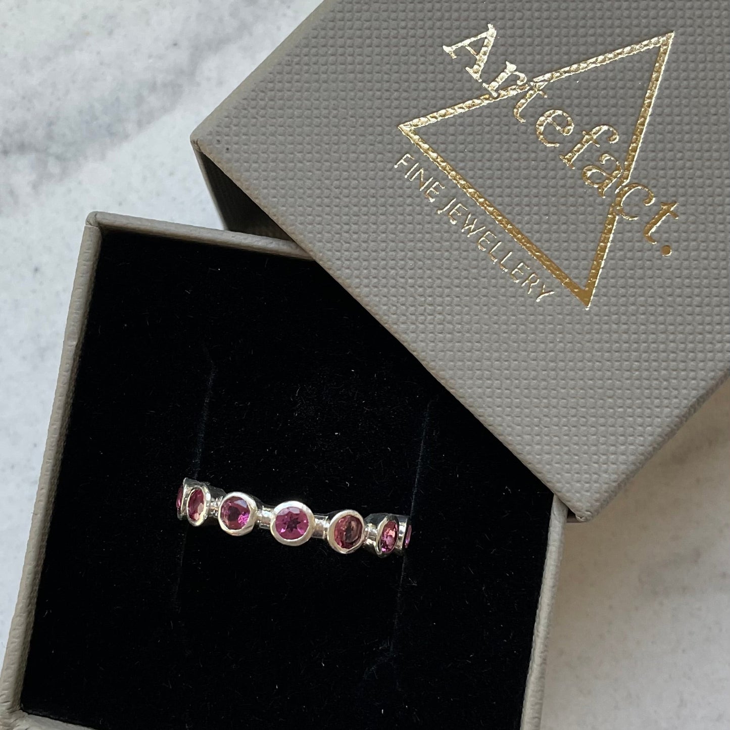 A silver band ring with five small round pink gemstones, presented in an open textured gray jewelry box with a black velvet interior. The lid of the box is propped behind it, showing the gold foil brand name 'Artfact Fine Jewellery'.