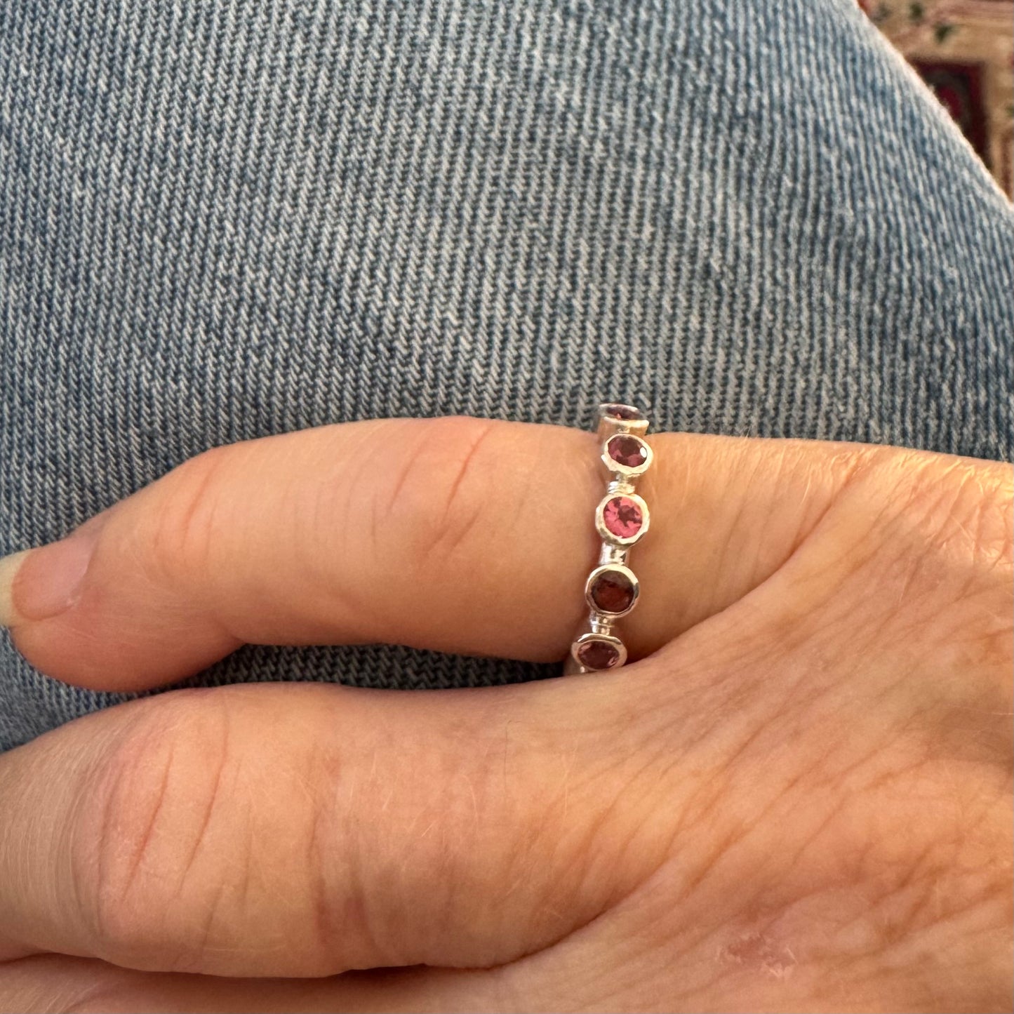 A close-up of a hand wearing a silver eternity ring with five round-cut pink gemstones, set against a background of blue denim fabric.